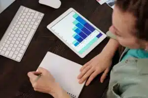 man writing on a notebook next to a tablet showing graphs